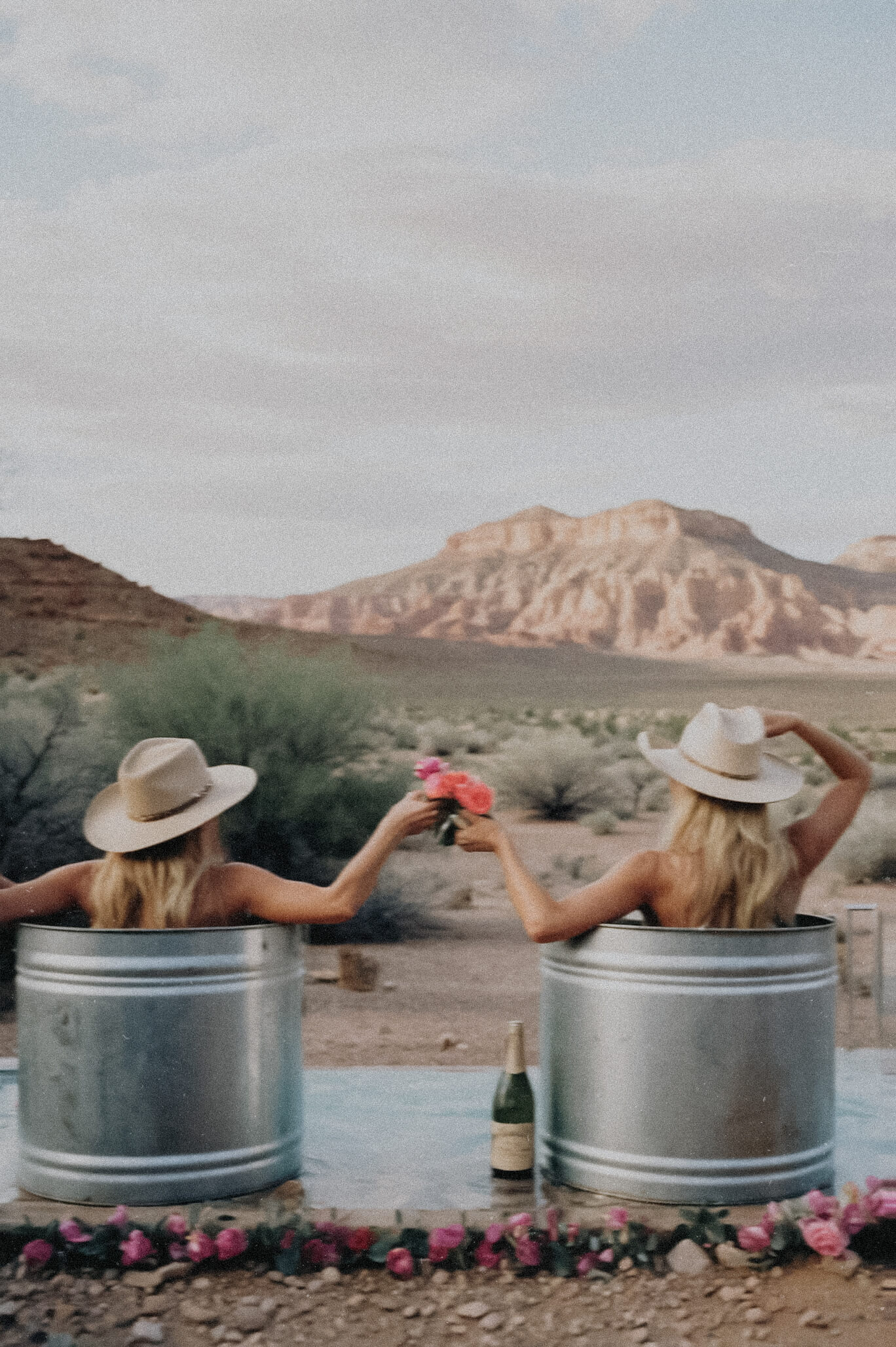Two women in cowboy hats sit in metal tubs outdoors, holding flowers and raising glasses, with desert mountains and a champagne bottle nearby—a perfect moment captured by Sweet Pea Ink Creative, experts in brand design.