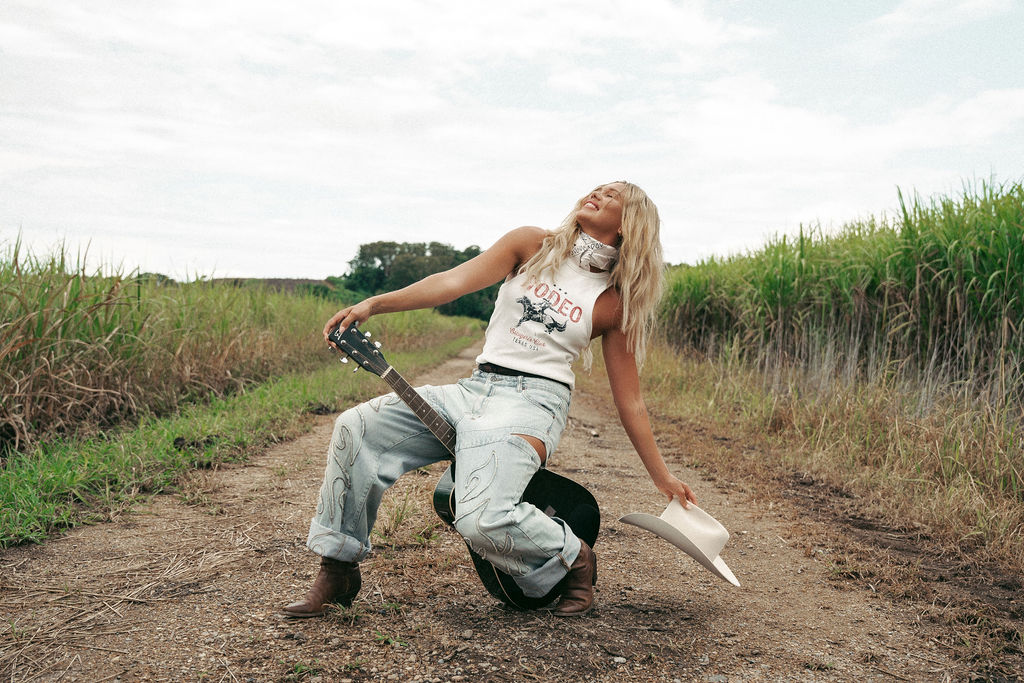 A country artist expresses joy while seated on a dirt road, holding a guitar. Tall grass frames the setting under an overcast sky, creating a tranquil ambiance, perfectly representing the brand identity of a client for Sweet Pea Ink Creative.